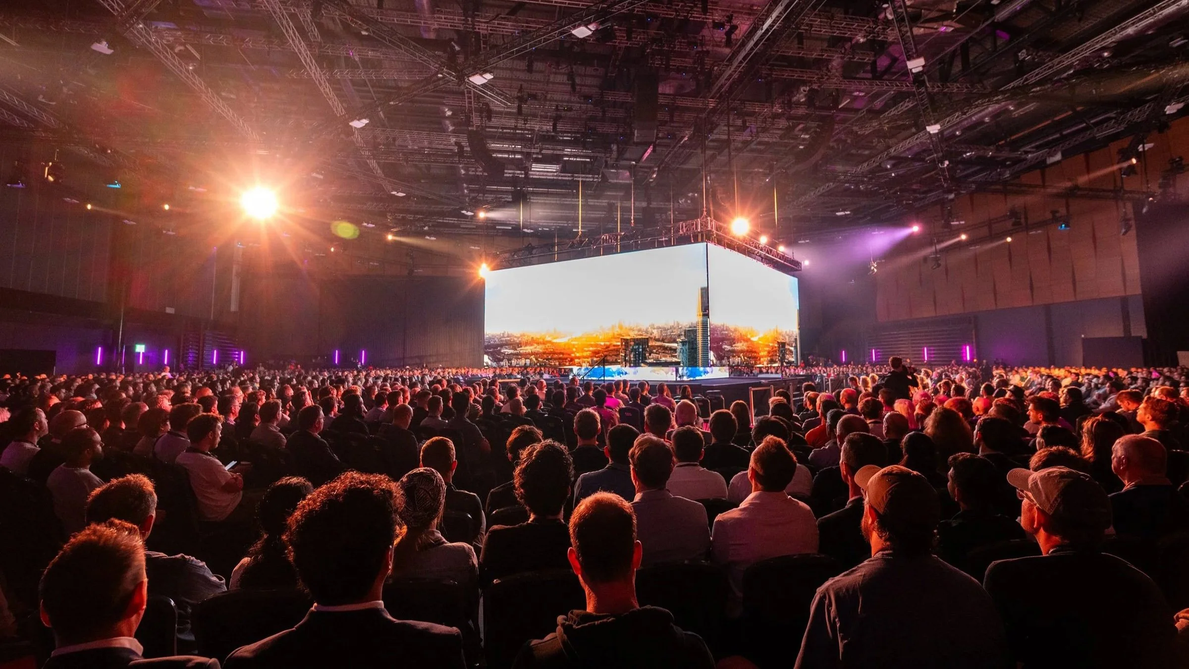 Large audience seated in a darkened conference or event hall, watching a giant screen displaying a city skyline at sunset, with stage lighting illuminating the scene.