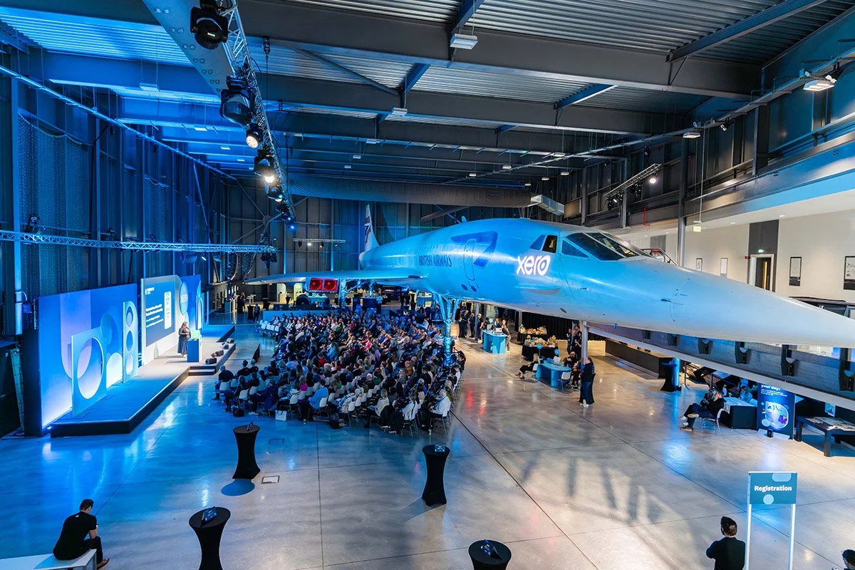 An indoor event space hosting a presentation with a large crowd seated and a speaker on stage. A spacecraft is on display behind the audience, marked with the Xero logo and UK flag.