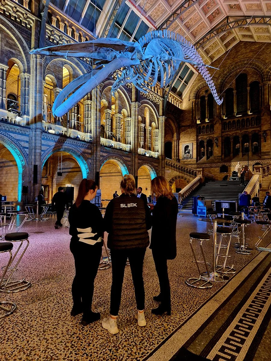 Three women are standing and talking in a grand, historic indoor space with a large whale skeleton hanging from the ceiling. The foyer has stone arches, ornate decorations, and a staircase in the background.