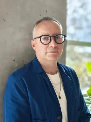A man with glasses, wearing a blue jacket over a white shirt, standing indoors near a concrete wall with a window showing a blurred outdoor scene.
