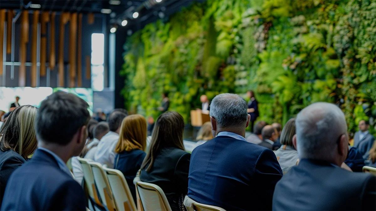 An audience attending a conference or seminar in a room with a green wall and wood accents.