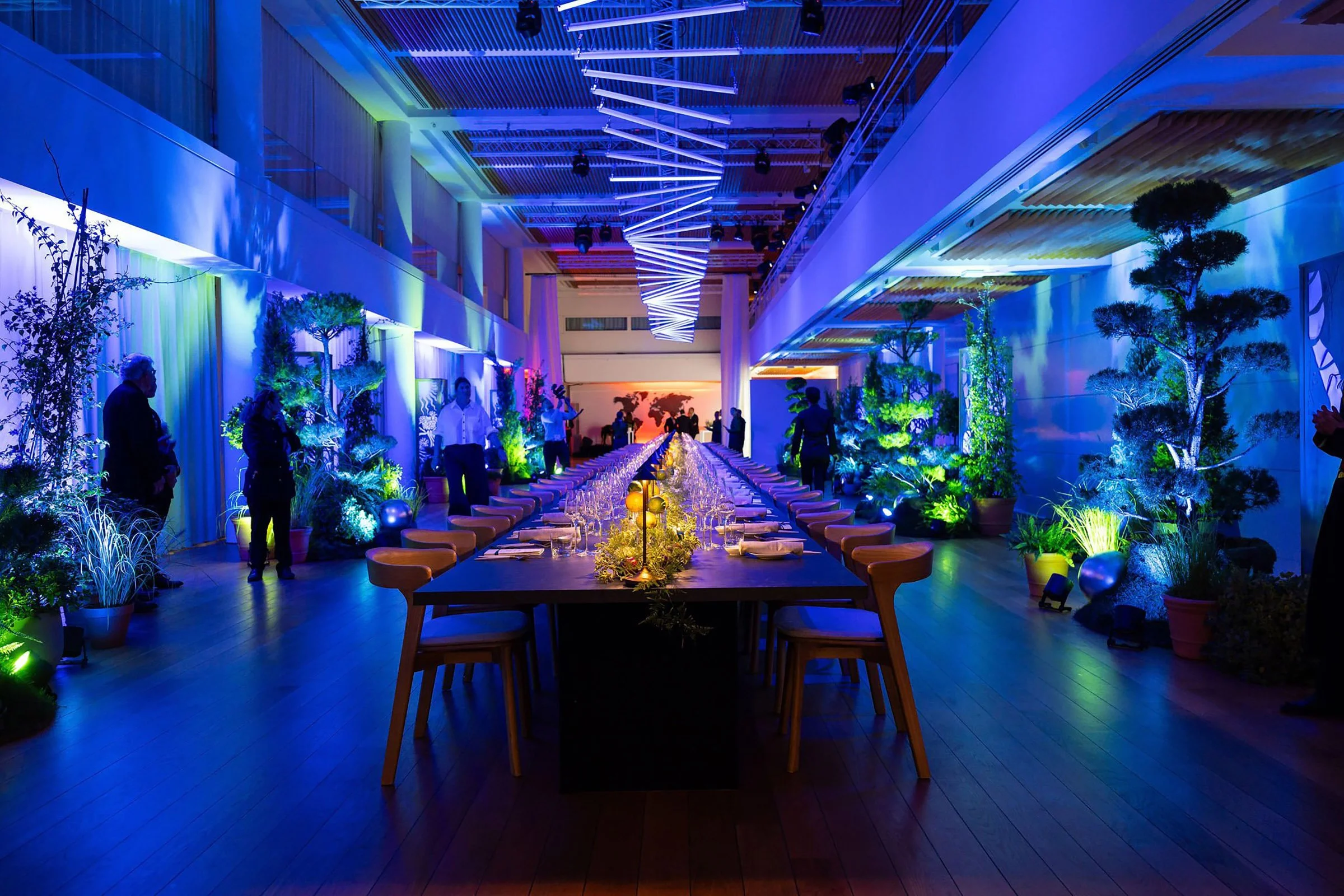 A large event space decorated with colorful lighting, potted plants, and a long table set for a banquet with glasses, plates, and floral centerpieces.