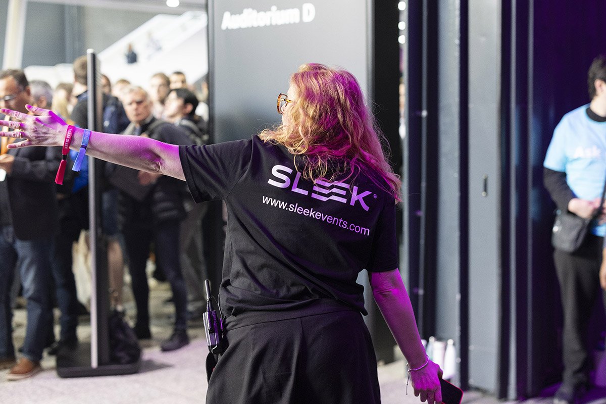 A woman with long, wavy hair wearing glasses, a black shirt with the words "SLEEK" and a website URL, standing with her back to the camera and extending her arm, in a crowded indoor event or conference.
