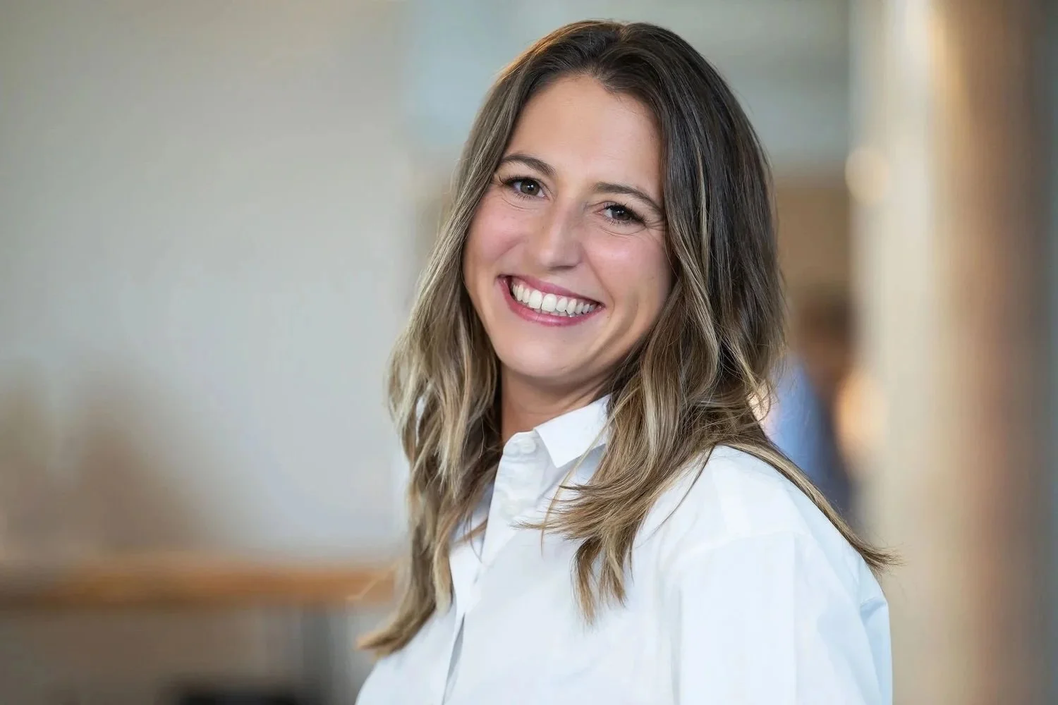 A woman with shoulder-length brown hair and light eyes smiling, wearing a white shirt in an indoor setting.