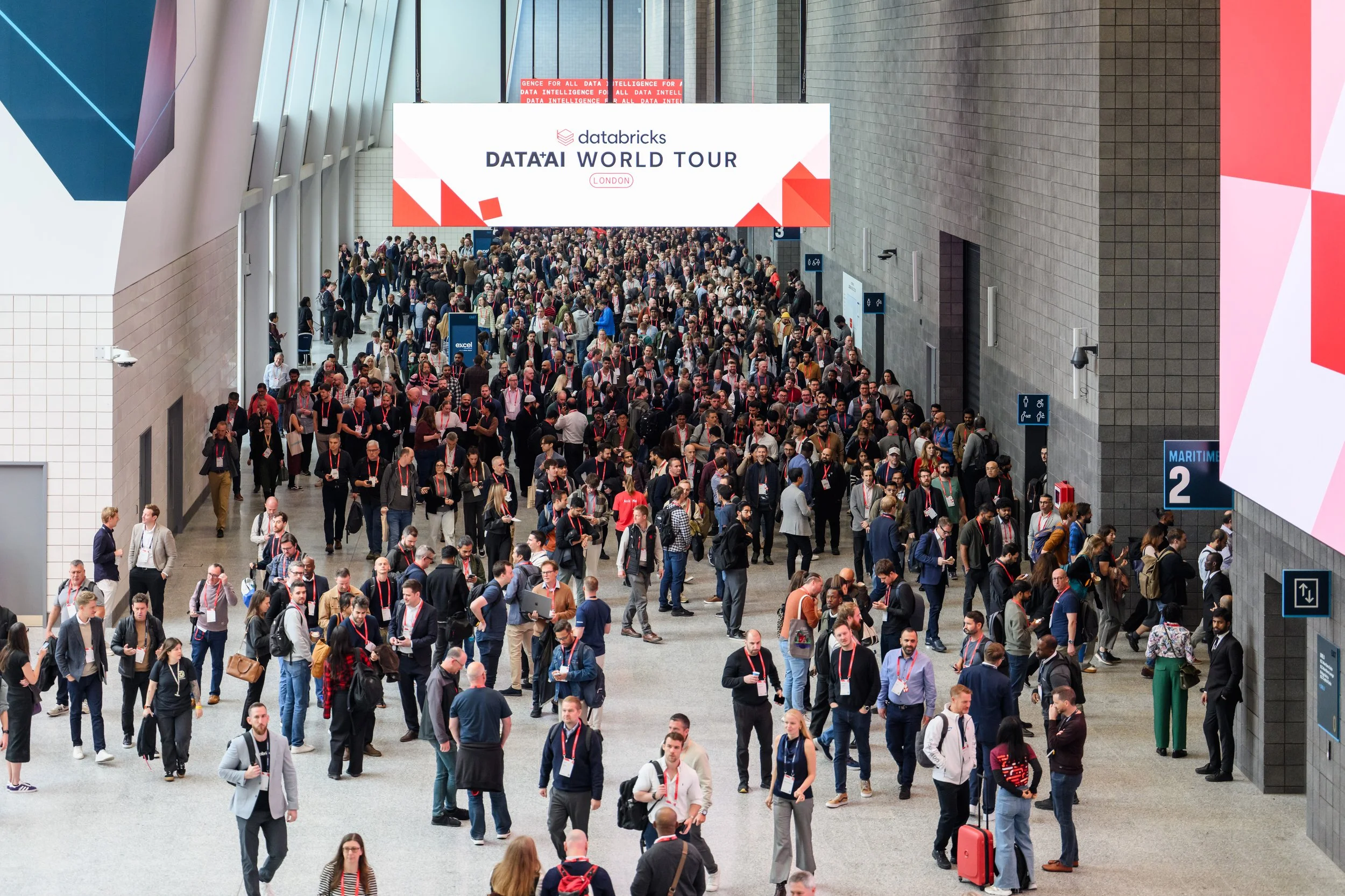 Large crowd of people walking through an indoor conference or event space with digital signage overhead displaying 'Data+AI World Tour London'.