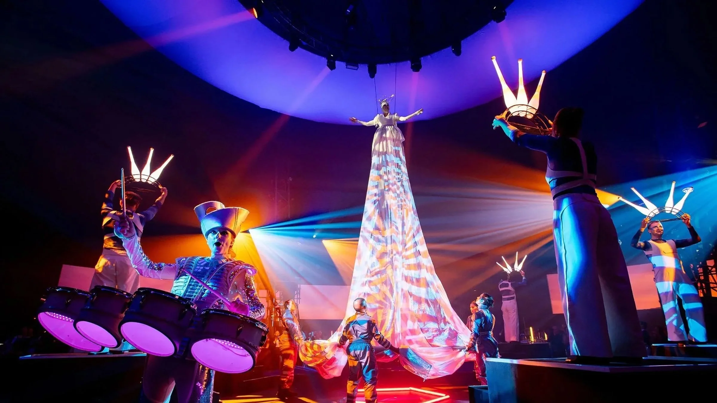 A theatrical performance with multiple performers on stage, featuring colorful costumes, dramatic lighting, and a central figure on a tall, illuminated structure.