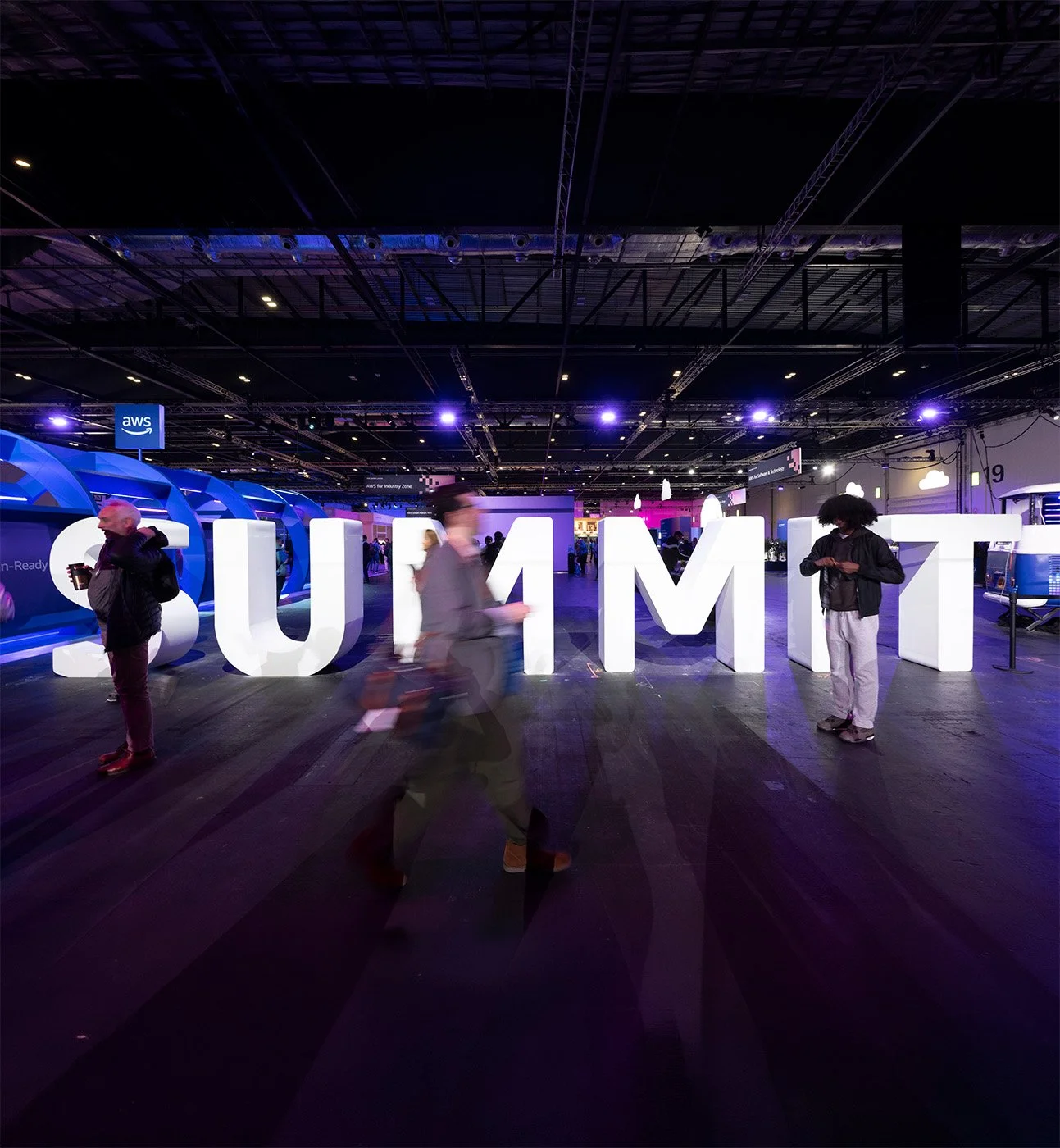 Large illuminated letters spelling 'SUMMIT' at a technology conference, with people walking past in a dimly lit exhibition hall.