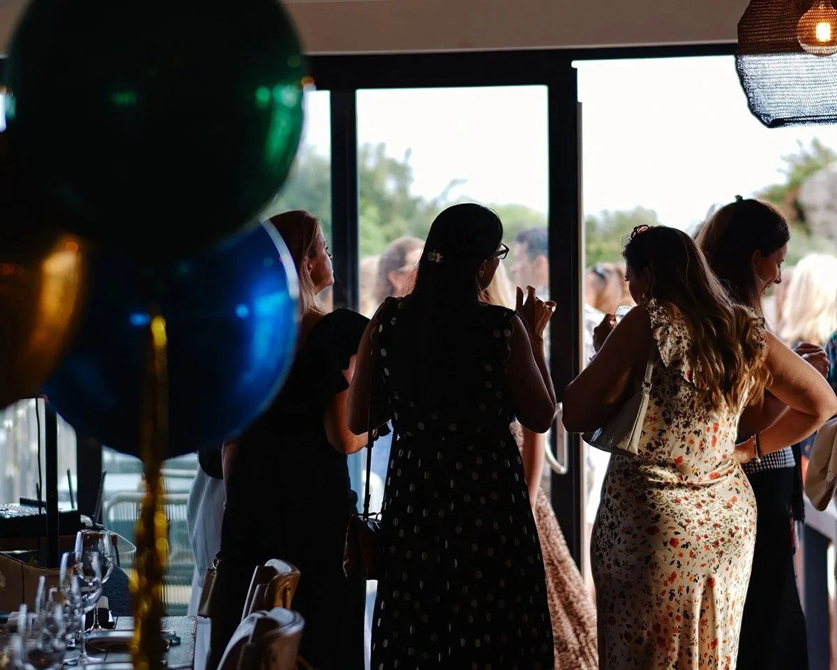 Group of women at a social gathering, standing near large windows with a view outside, some talking and others looking at their phones, with balloons in the foreground.