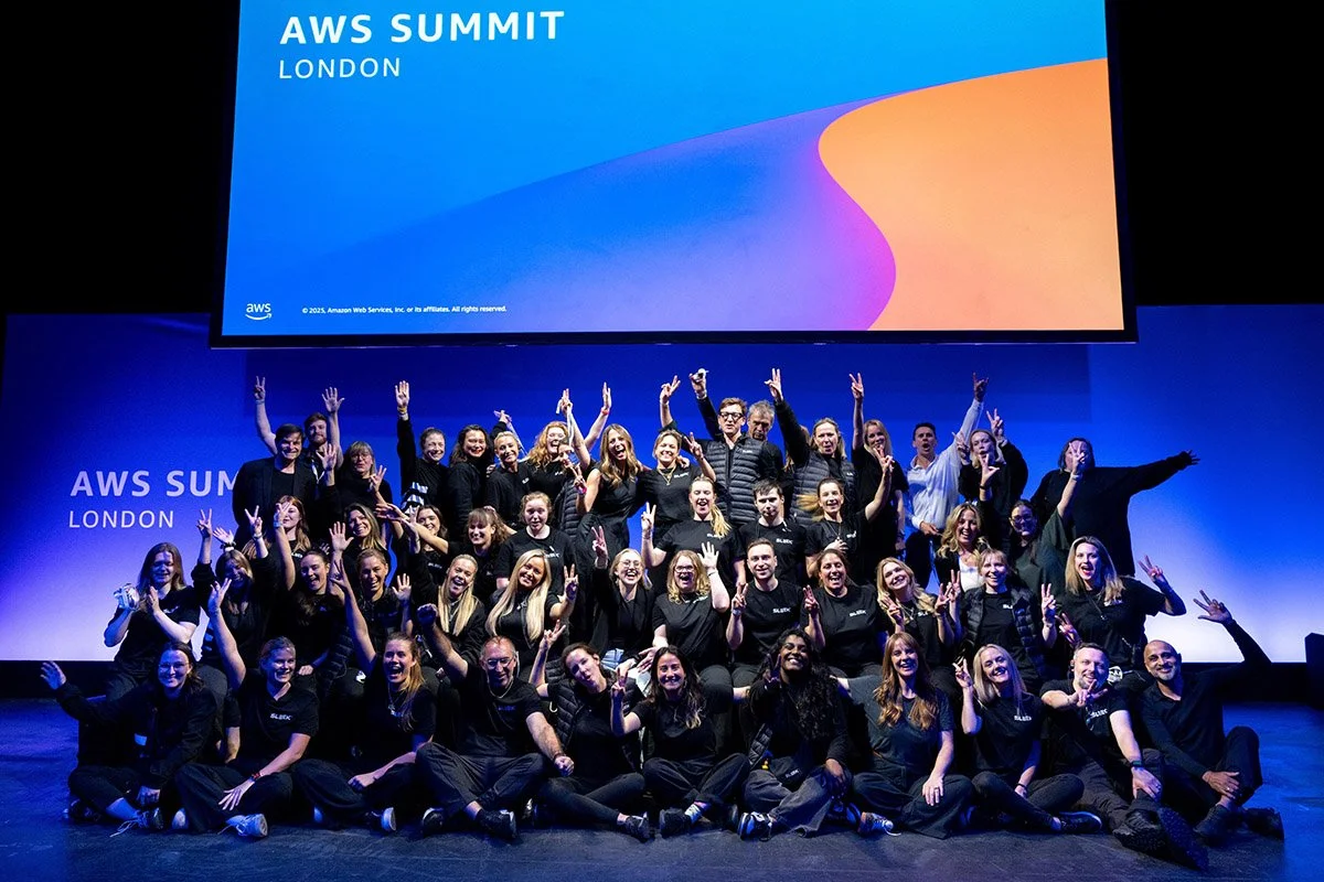 Group of people on stage at AWS Summit London, posing with peace signs and smiling in front of a large screen displaying the AWS Summit London logo.