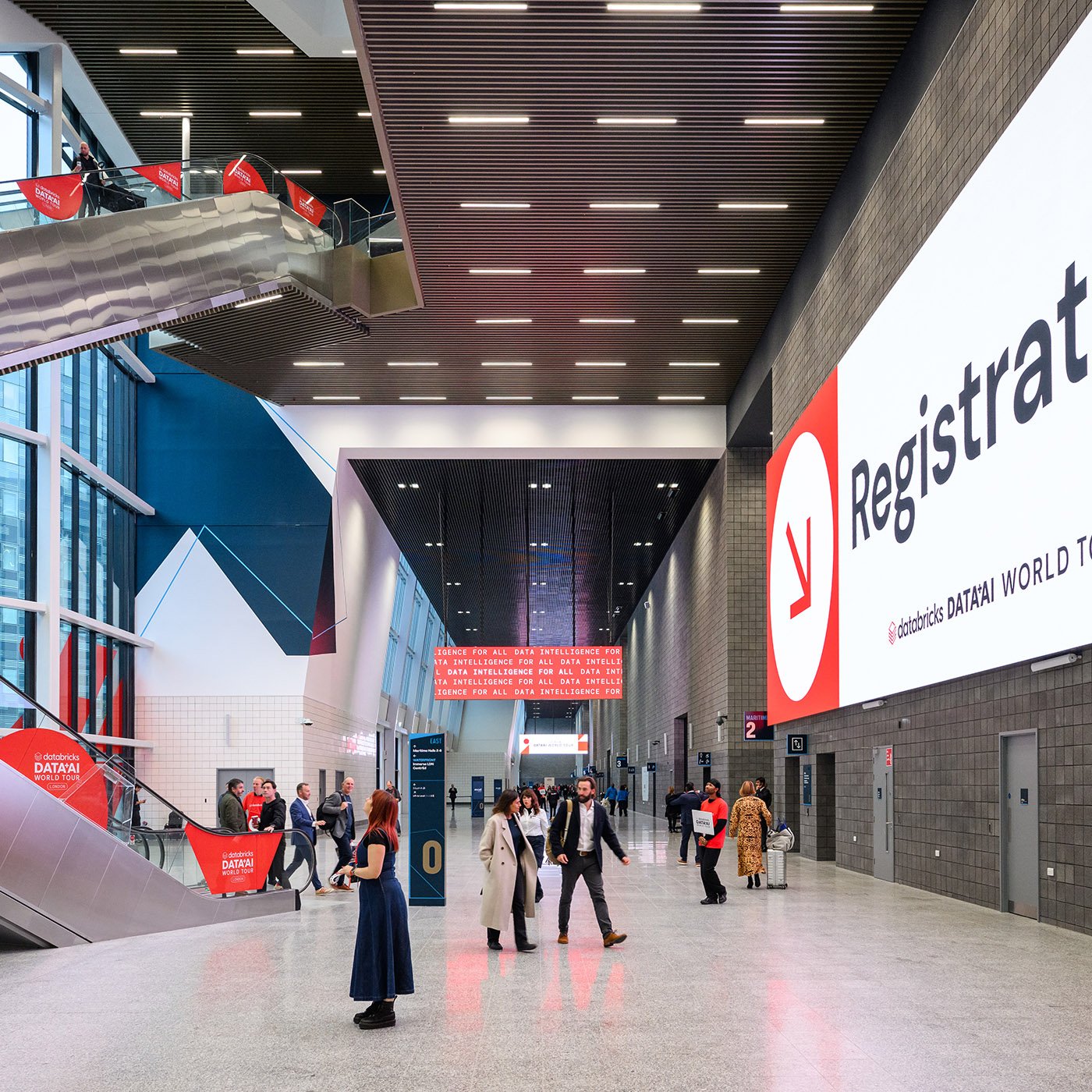 Interior of a modern conference center with a large digital registration sign, people walking and talking, escalators, and informational signs.