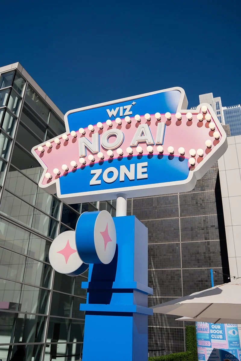 A sign with a pink and blue color scheme reading 'WIZ NO A! ZONE' with lit bulbs around the edges, situated in an urban area with glass buildings and a blue sky in the background.