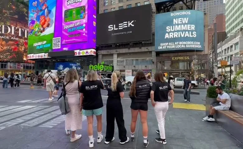 Five women standing on a city sidewalk, with billboards and digital screens behind them, Times Square in New York City.