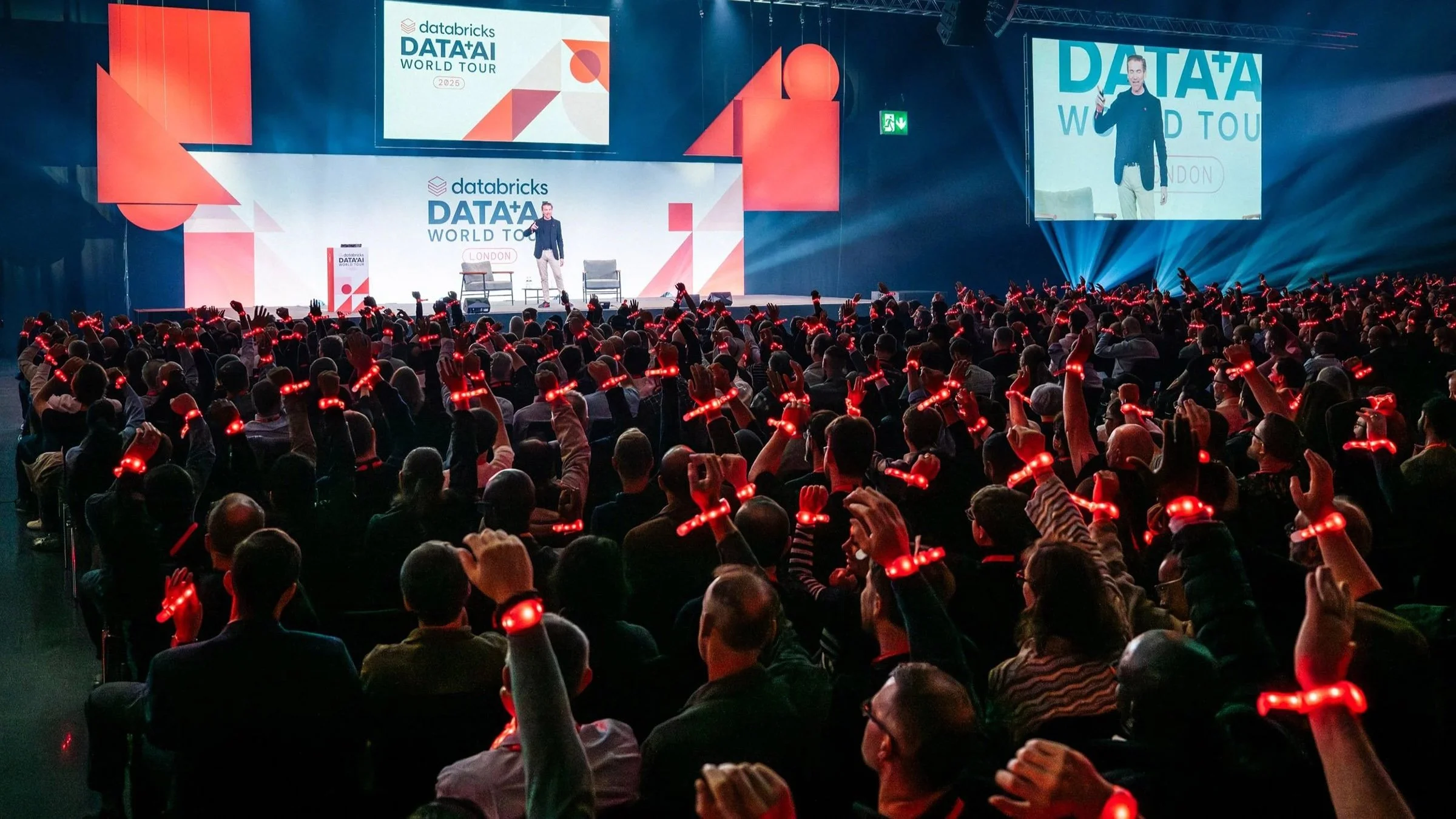 A large crowd attending a conference or event on data and AI in a spacious auditorium. The stage features a speaker presenting with a backdrop displaying 'databricks DATA+AI WORLD TOUR 2025' and large screens projecting the speaker and presentation. Audience members are wearing red LED gloves, raising their hands, and listening attentively.