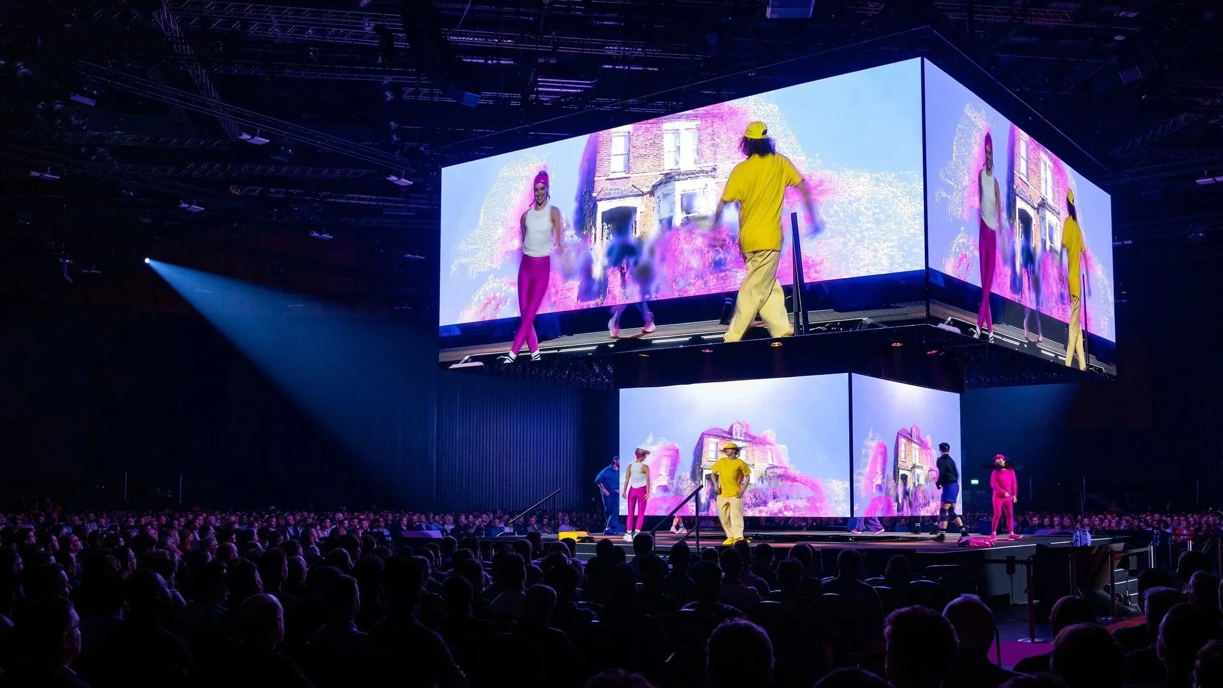 A stage with vibrant digital screens displaying artistic images, performers dressed in colorful casual clothing, and a large audience watching a live event in a darkened arena.