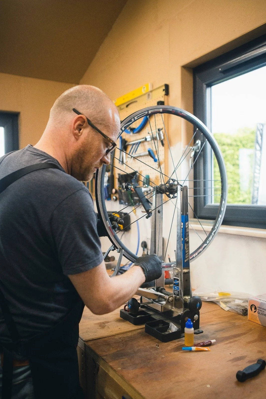 A person working on a bicycle wheel mounted on a bike repair stand in a workshop, with tools and supplies on the workbench.