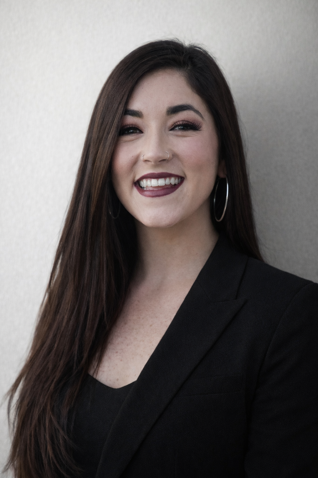 A smiling woman with long dark brown hair, hoop earrings, and a nose ring, wearing a black blazer against a neutral background.