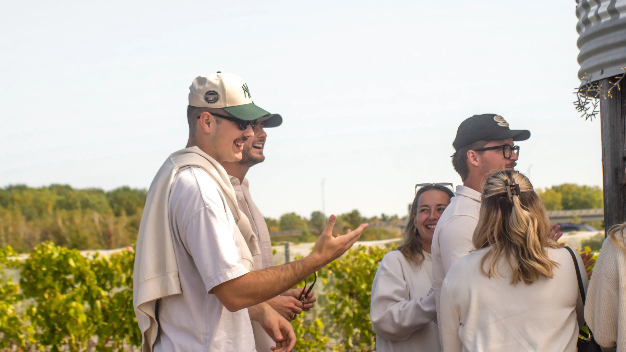 Groupe de personnes souriantes discutant en extérieur, portant des vêtements blancs, avec un paysage verdoyant en arrière-plan. Pris en photo à côté du silo de Vineterra