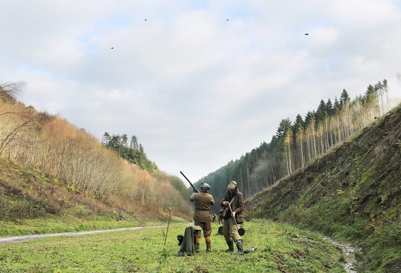 Two people dressed in outdoor clothing, wearing helmets, standing in a grassy area between hills with trees, looking at and discussing something, with remote controls on the ground, and helicopters flying in the sky above.
