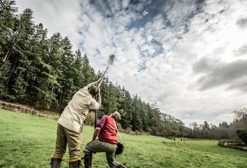 Two people, a man and a woman, are in a grassy field with trees in the background. The man is holding a long pole or tool, and the woman is crouched down, both looking up at the sky where there is a patch of clouds.