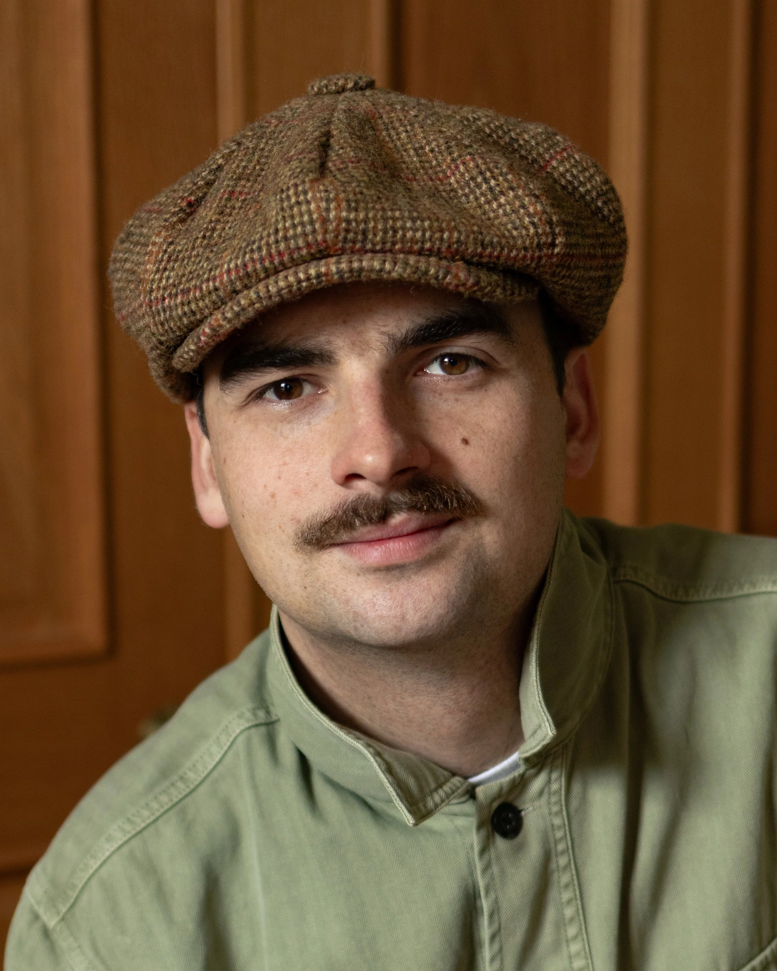 Close-up photo of a man's face, focusing on his eyes, nose, and mouth, wearing a brown herringbone flat cap and a light green collared shirt with a button, with a wooden background.