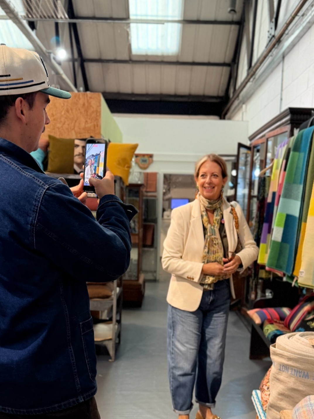 A woman with short hair, wearing a white blazer, a patterned scarf, and blue jeans, is smiling while posing for a photo in an indoor store that sells textiles and home decor.