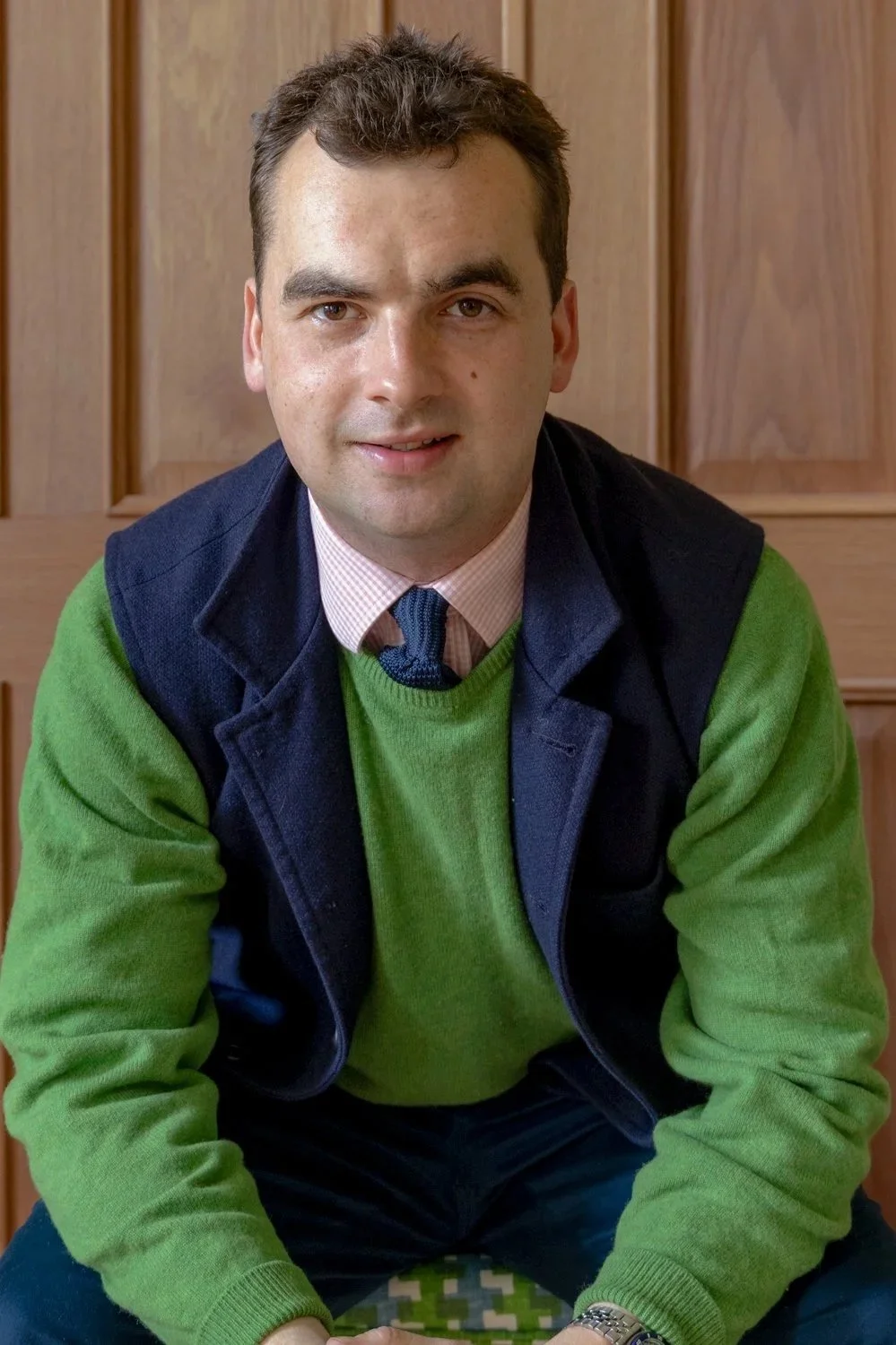 A man with short dark hair, wearing a green sweater over a collared shirt and tie, sitting in front of a wooden panel wall.