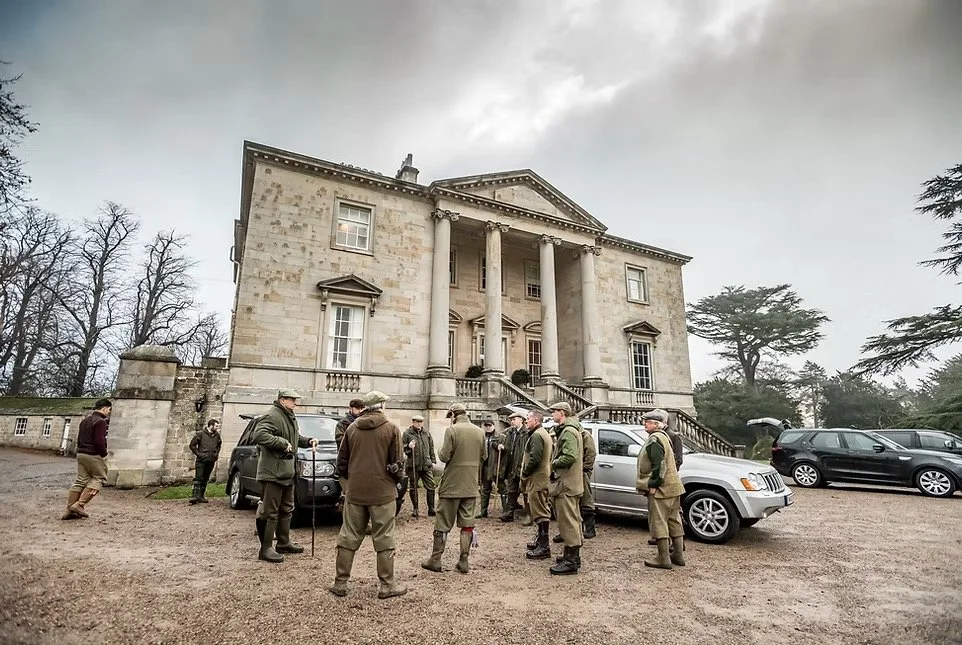 Group of men in hunting gear gathered outside a large historic mansion with columns and stairs, cloudy sky in the background.