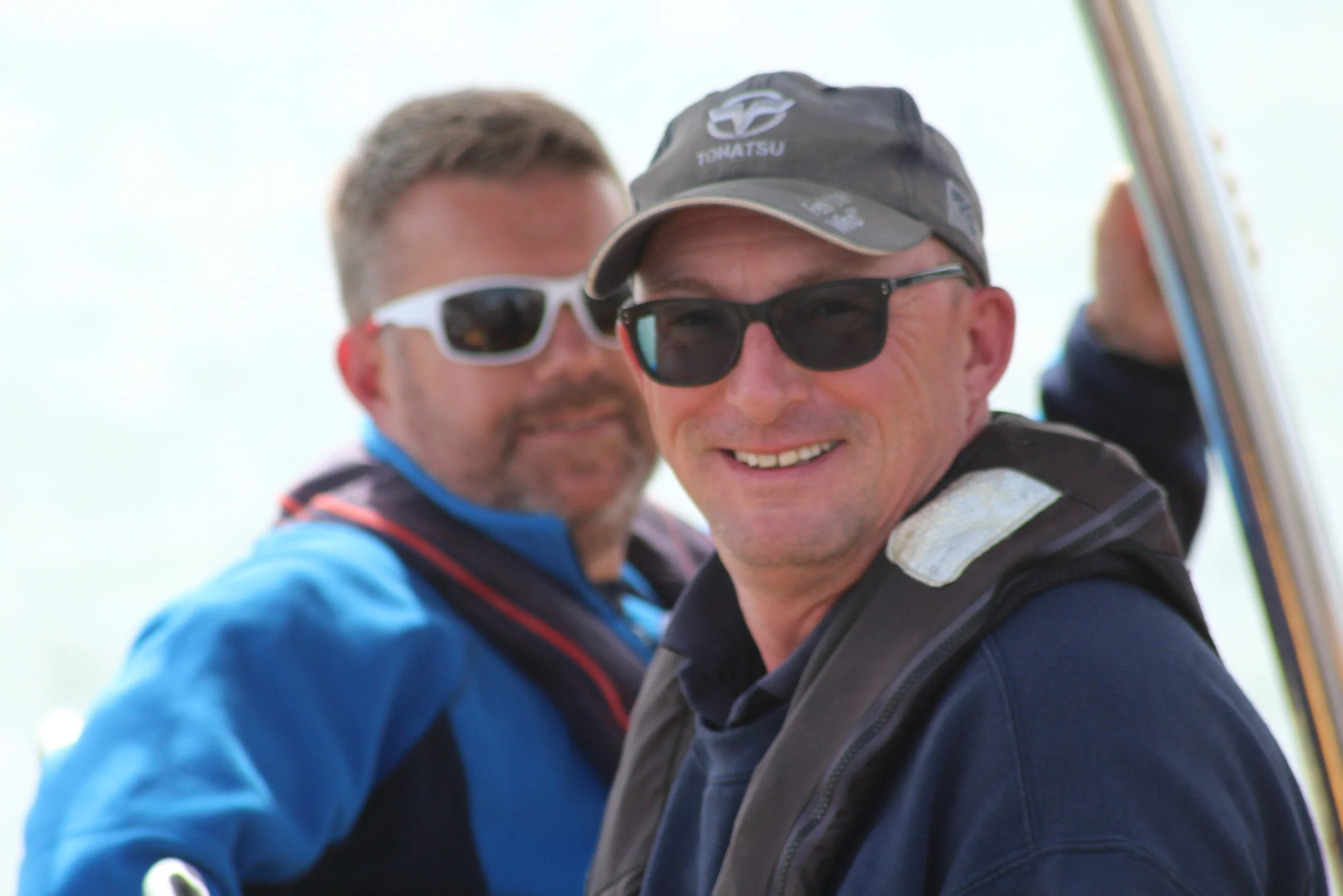Smiling man wearing sunglasses, a gray cap, and a black jacket outdoors.