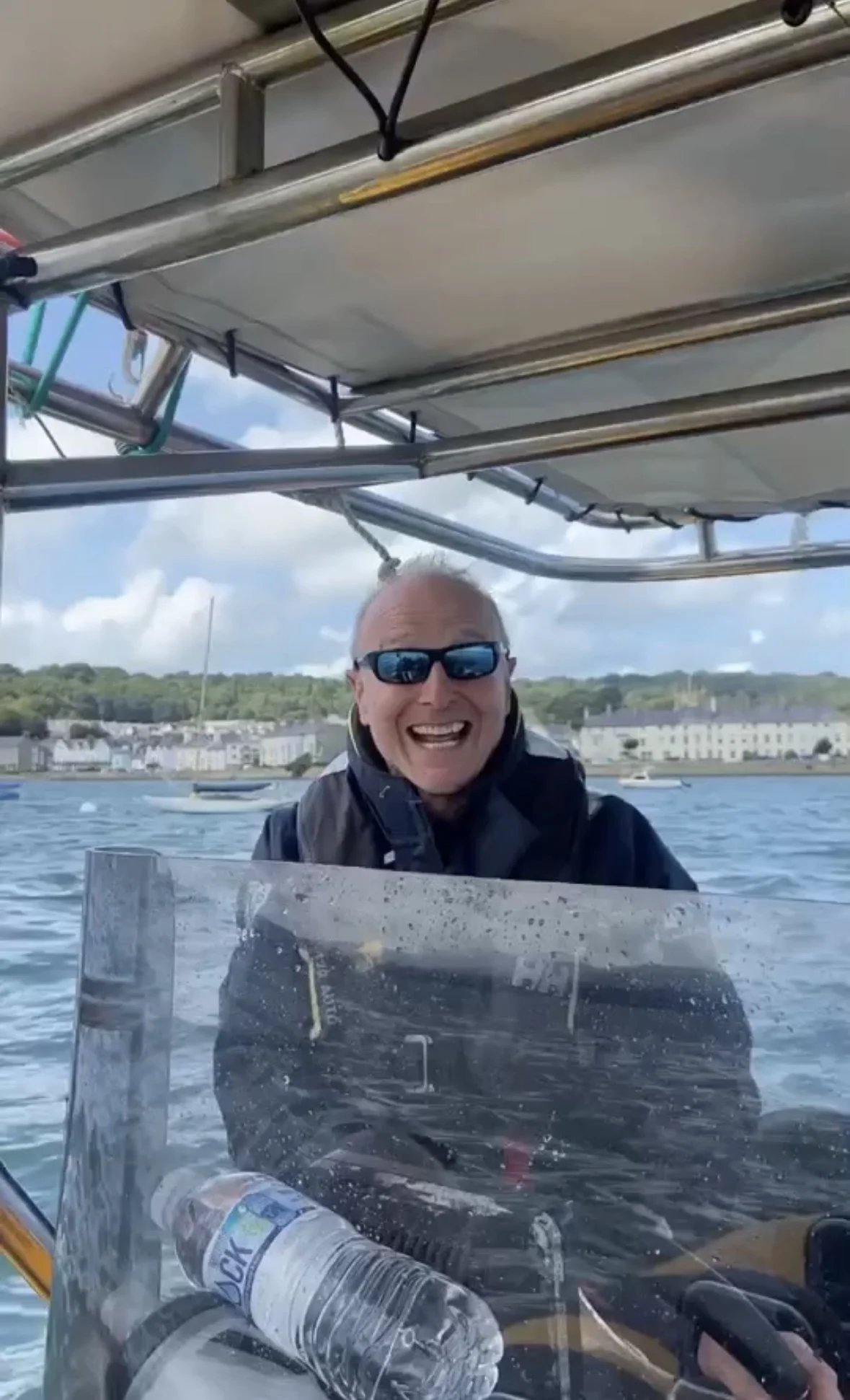 Man wearing sunglasses, a black beanie, and life jacket on a boat with water and cloudy sky in the background.