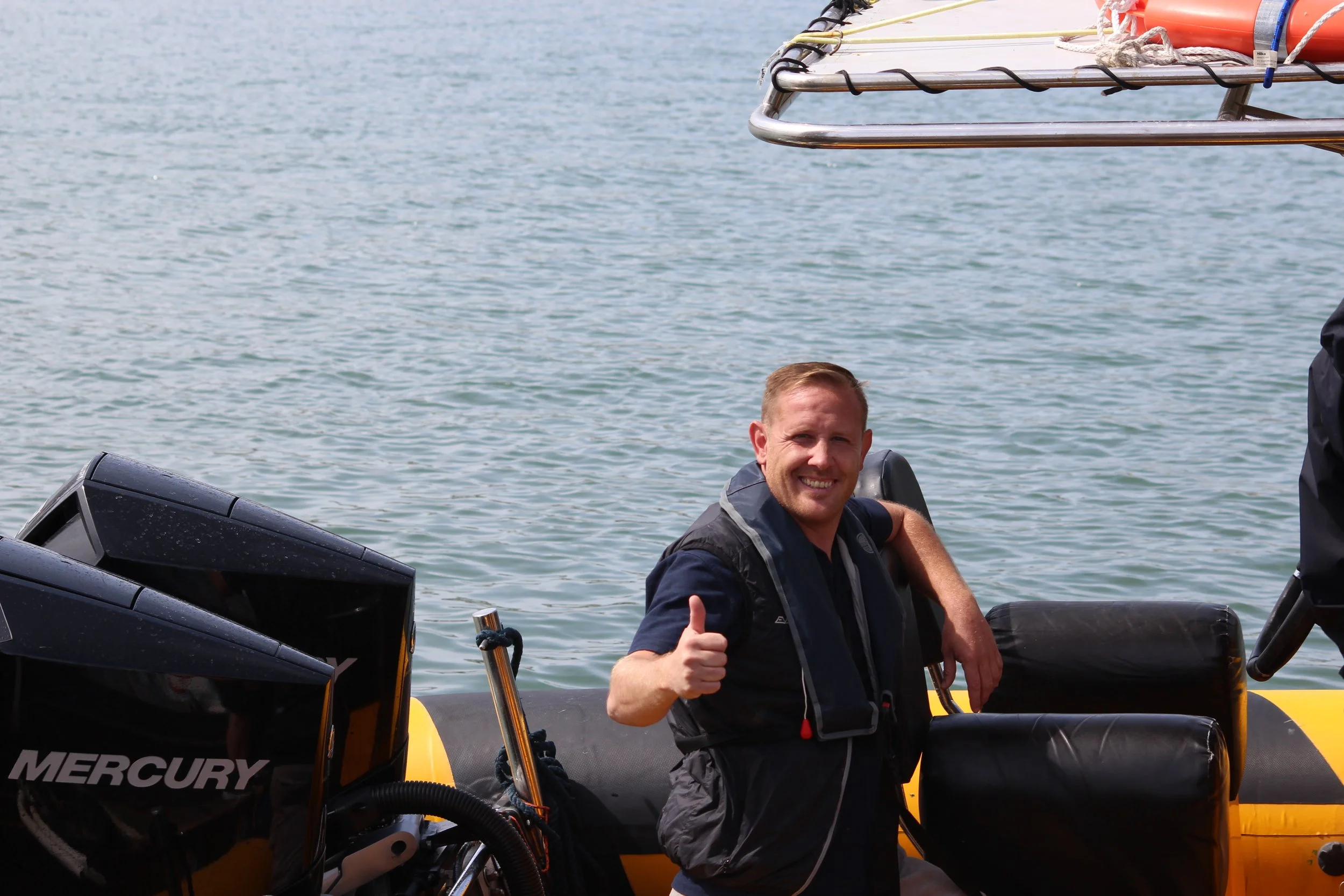 A smiling man with short blond hair outdoors near water, holding a paddle or oar.