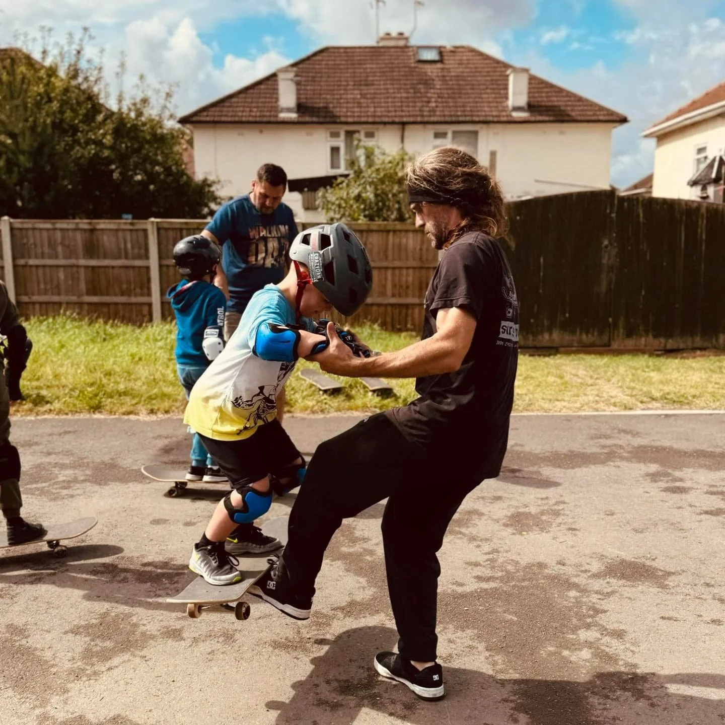 A man teaching young children how to skateboard outdoors on a sunny day. The children are wearing helmets, and one child is actively riding a skateboard while others wait nearby.