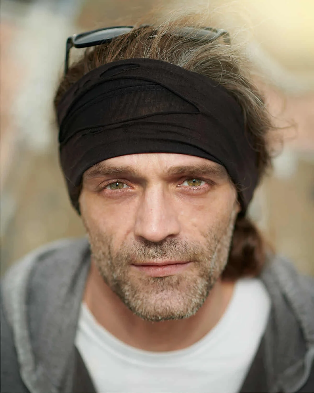 Close-up portrait of a man with gray hair, wearing a black headband, glasses on his head, and a gray hoodie, looking directly at the camera.