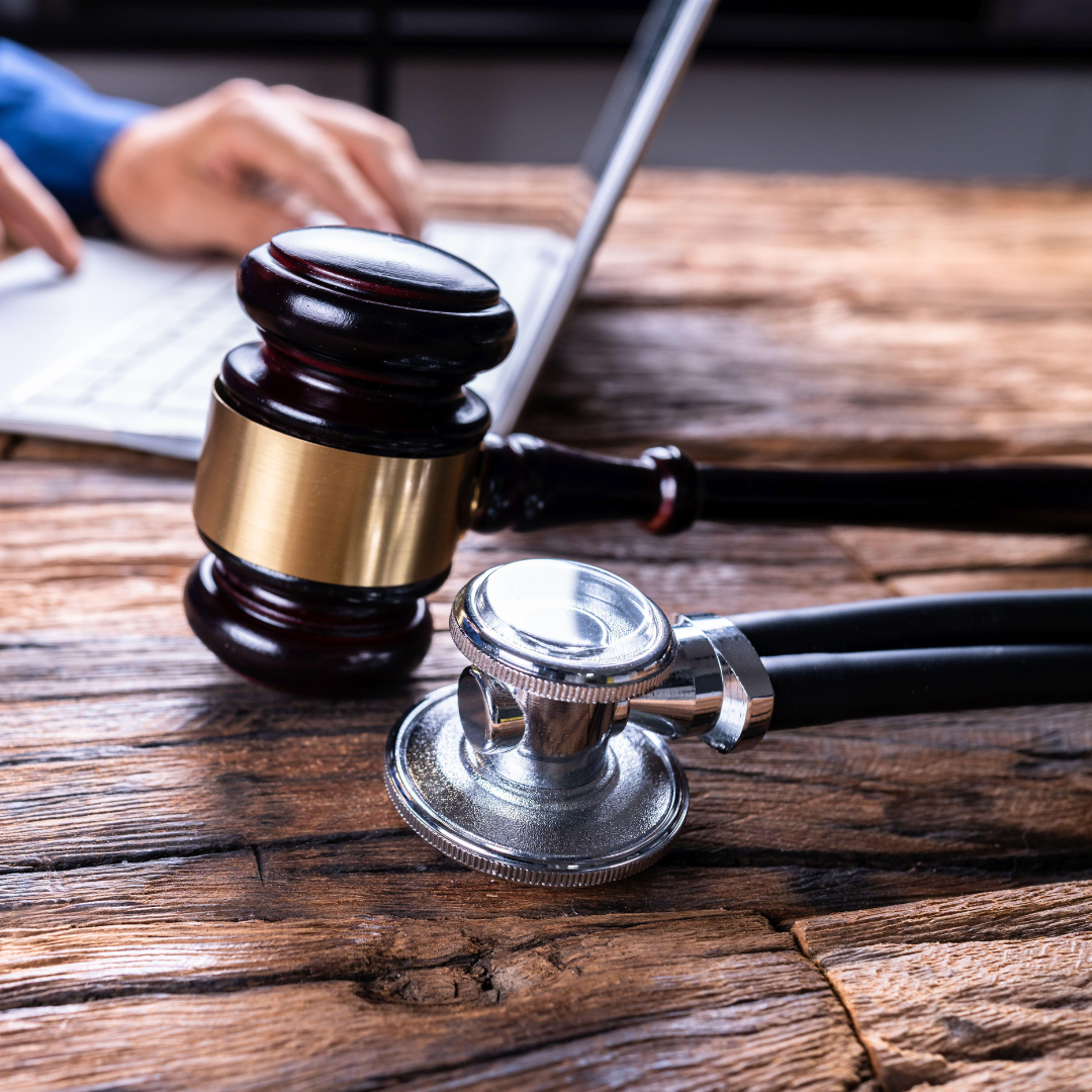A judge's gavel and a stethoscope on a wooden table, with a person typing on a laptop in the background.