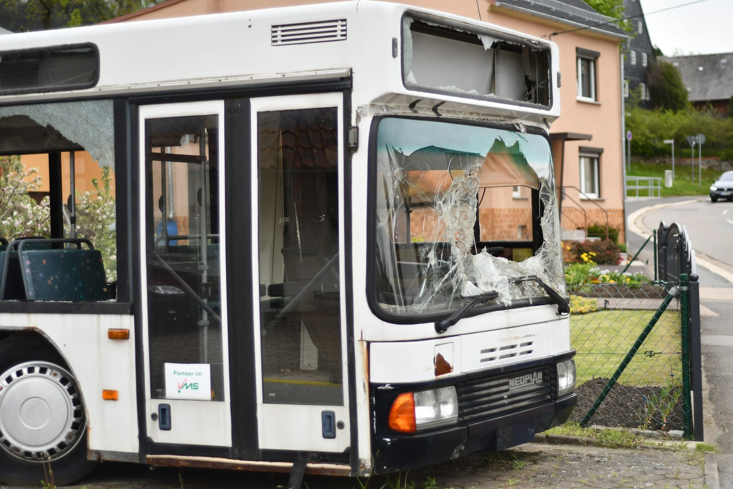 A white bus with a broken front windshield and shattered glass on the front window parked in a residential area.
