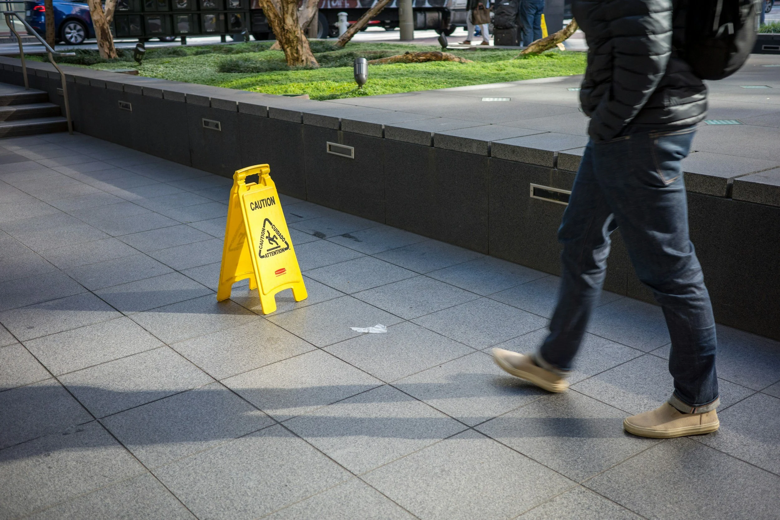 A person in dark clothing and tan shoes walking past a yellow caution sign on a sidewalk in an urban area. The sign warns about wet floor. There is a small landscaped area with trees and lights behind the sidewalk. Vehicles and people are visible in the background.