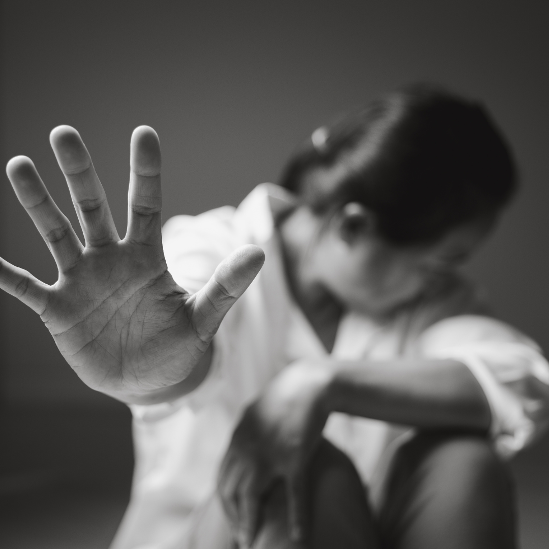 A person with long hair and a white shirt extends their arm, with their palm open and fingers spread, while sitting with their head down and eyes closed.