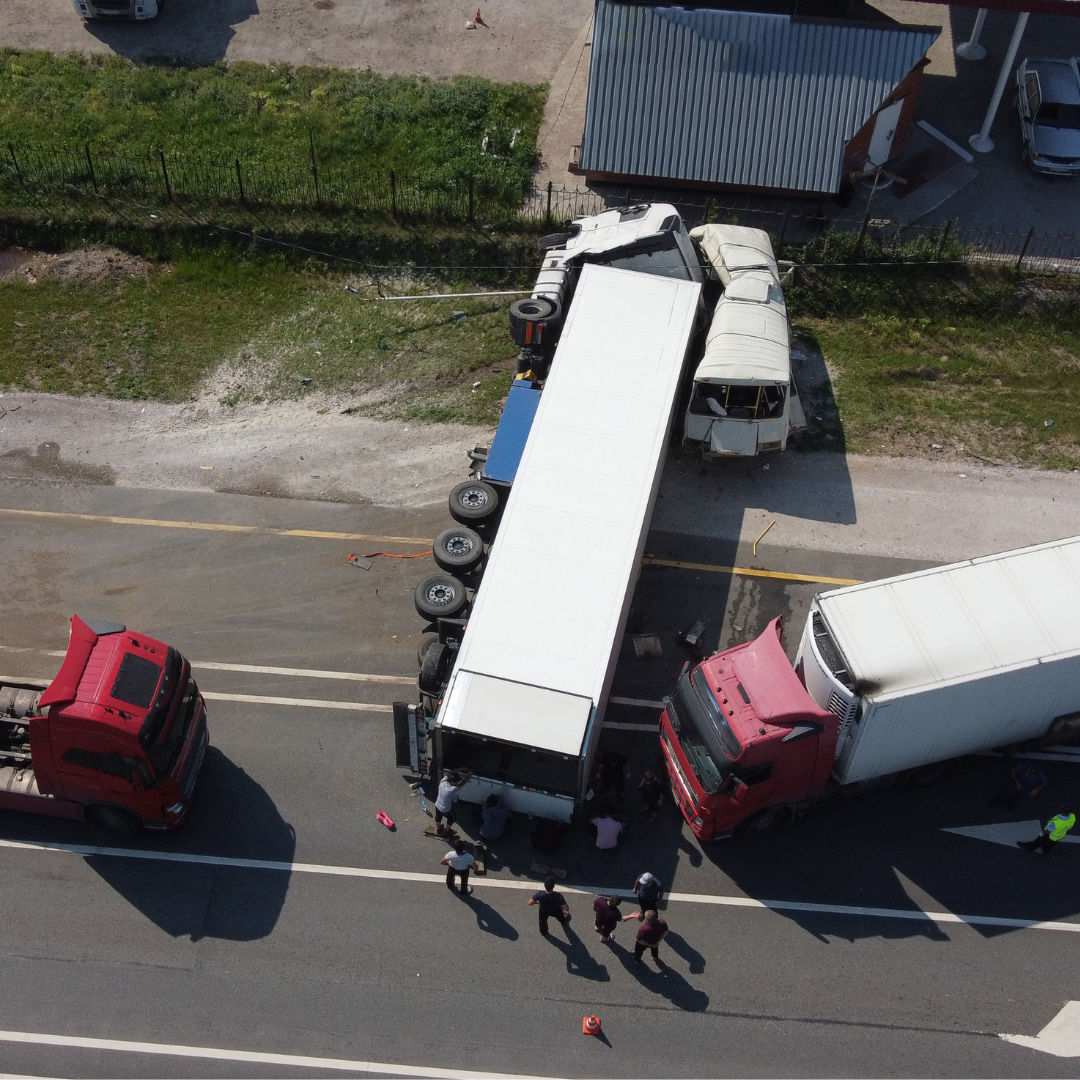 An aerial view of a multi-vehicle accident on a road, involving large trucks and a van. Several people are gathered around the scene, inspecting and possibly assisting. A damaged van is partly off the road, and the trucks are stopped nearby.