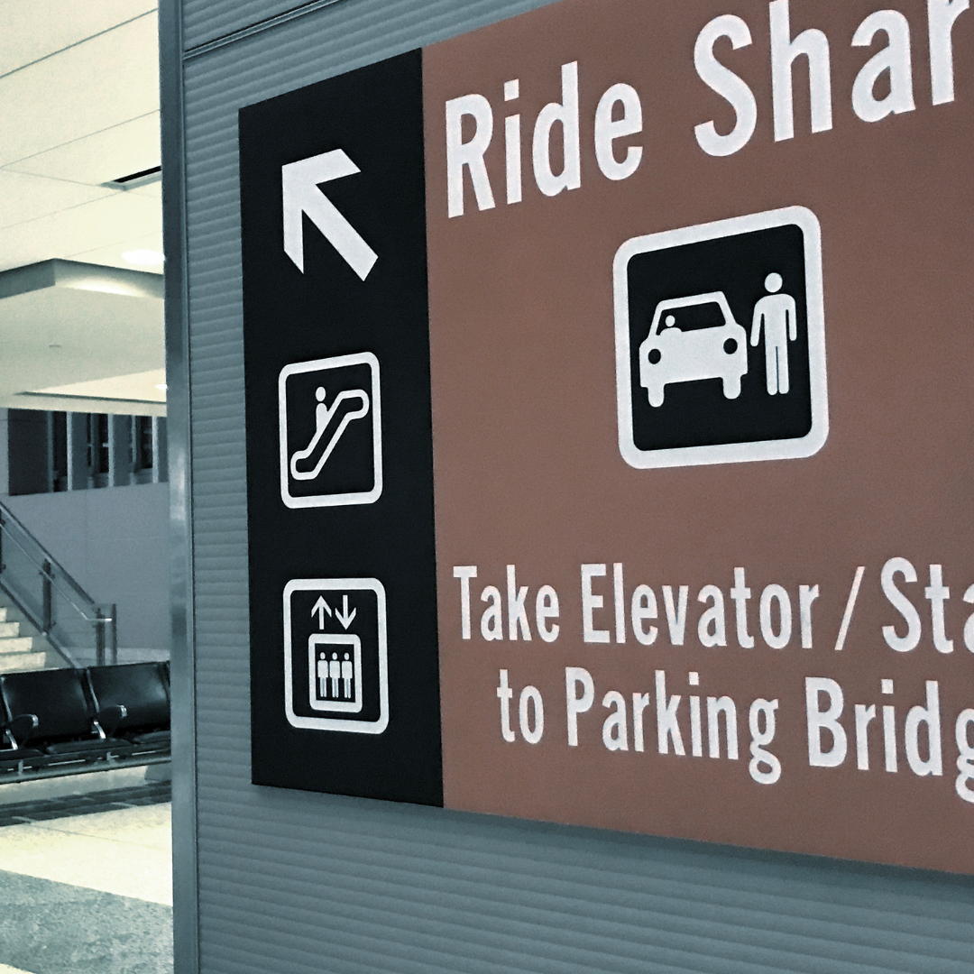Sign at an airport indicating directions to ride sharing, escalator, and elevator, with an icon for taking an elevator or stairs to the parking garage.