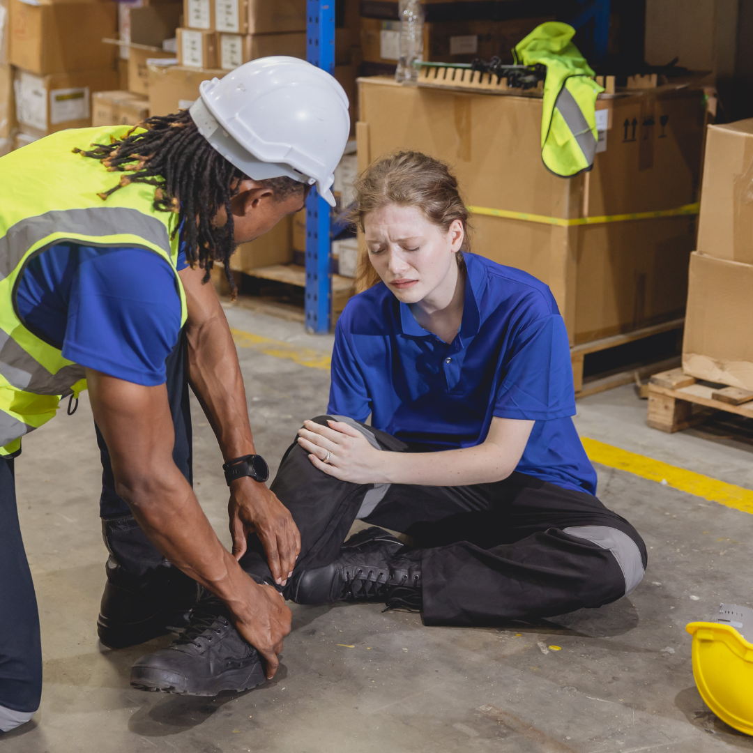 A worker in a yellow safety vest and white helmet helping a woman in a blue shirt who is sitting on the floor and appears to be injured in a warehouse with cardboard boxes and shelves in the background.