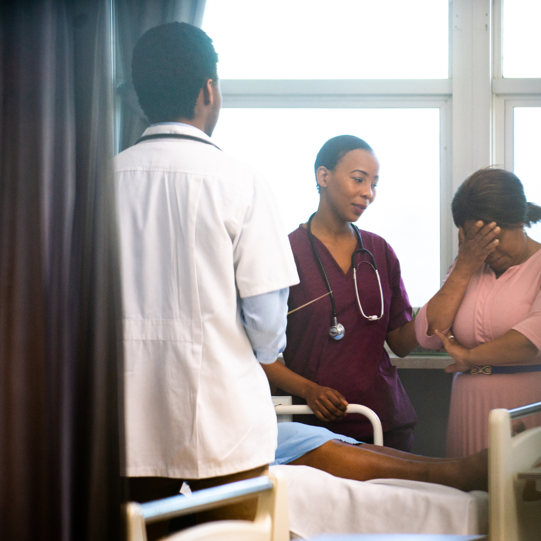 A nurse is comforting a woman in a hospital room, with another medical professional observing. The patient is lying in a hospital bed, and they are by a large window.