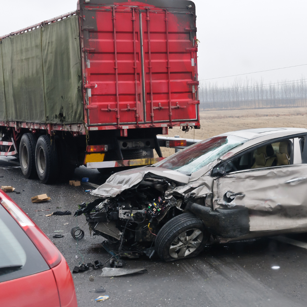 A car has been involved in a crash with a semi-truck on a highway. The front of the car is severely damaged, with parts and debris scattered on the road. The semi-truck appears to be stationary nearby.