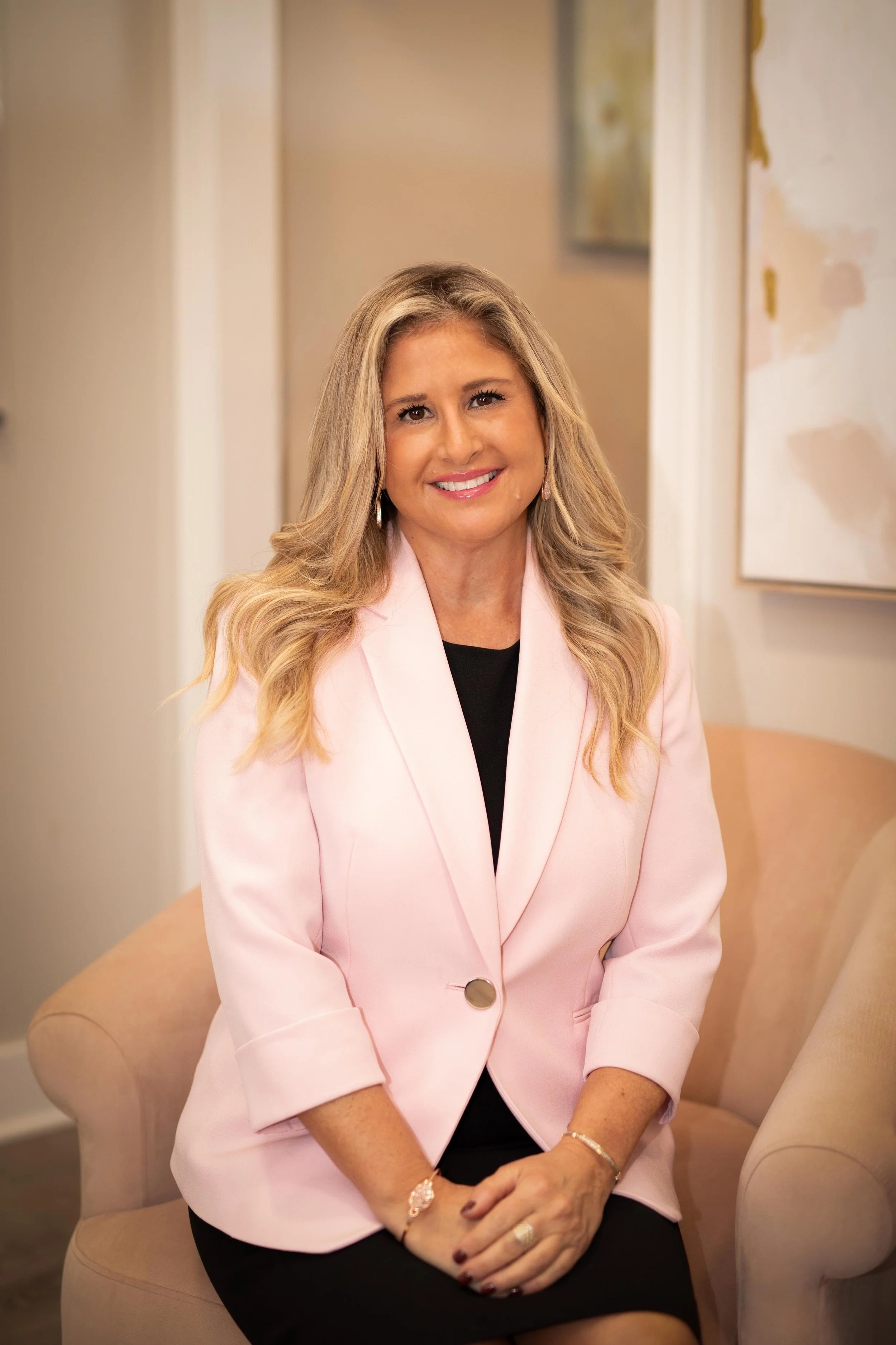 A smiling woman with long blonde hair sitting on a beige armchair in a light-colored room, wearing a pink blazer and black dress.