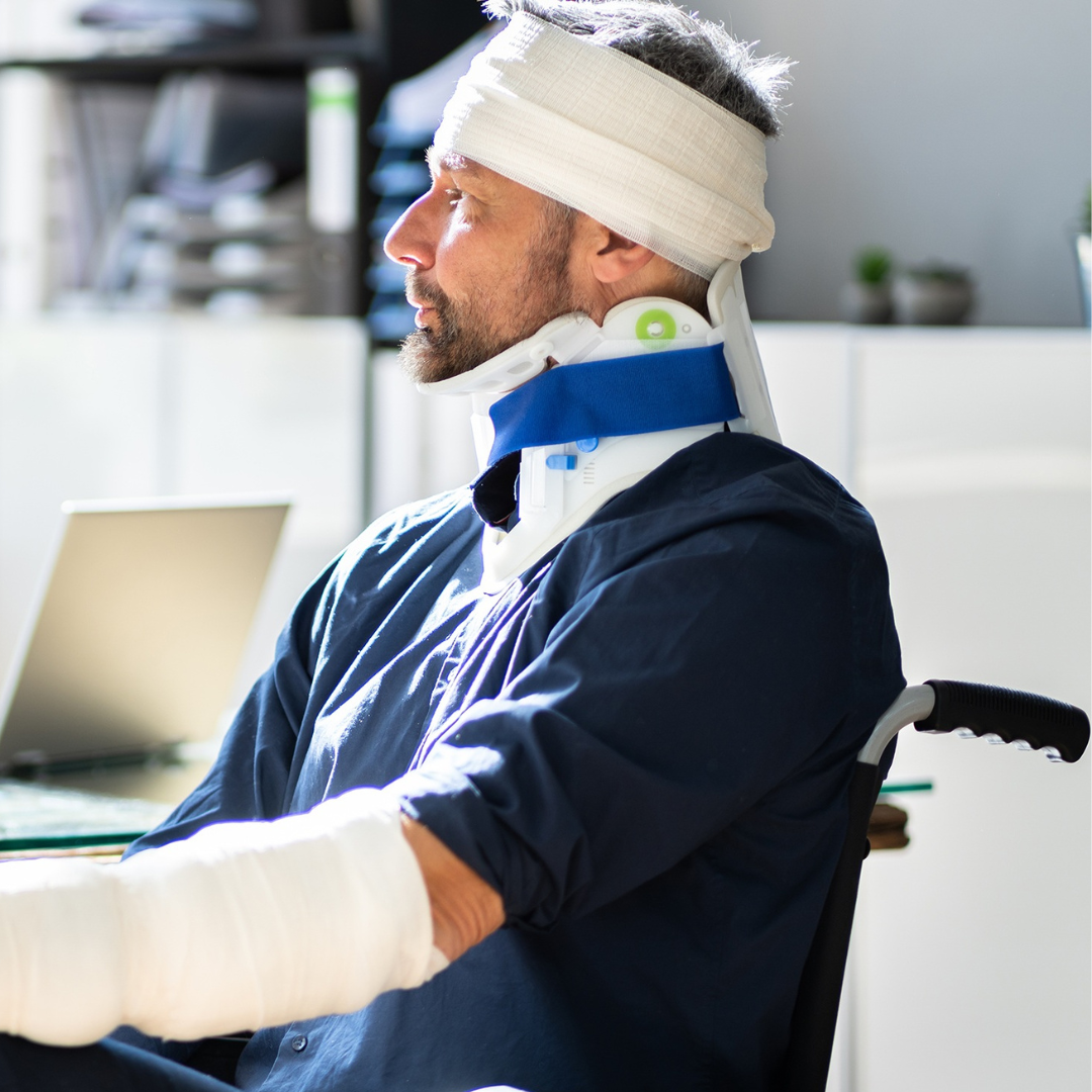 Man with bandaged head and neck, sitting in a wheelchair in an office, wearing a neck brace and a dark blue shirt.