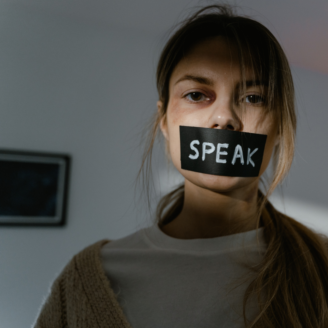 A young woman with brown hair and a neutral expression, has a black tape covering her mouth with the word 'SPEAK' written in white.