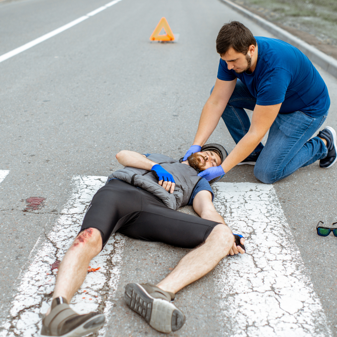A rescue worker attends to a man who appears to be injured and lying on a road with visible blood and a scrape on his leg. The rescuer is holding the man's head gently, and a warning triangle is placed on the road behind them, indicating an accident or emergency situation.
