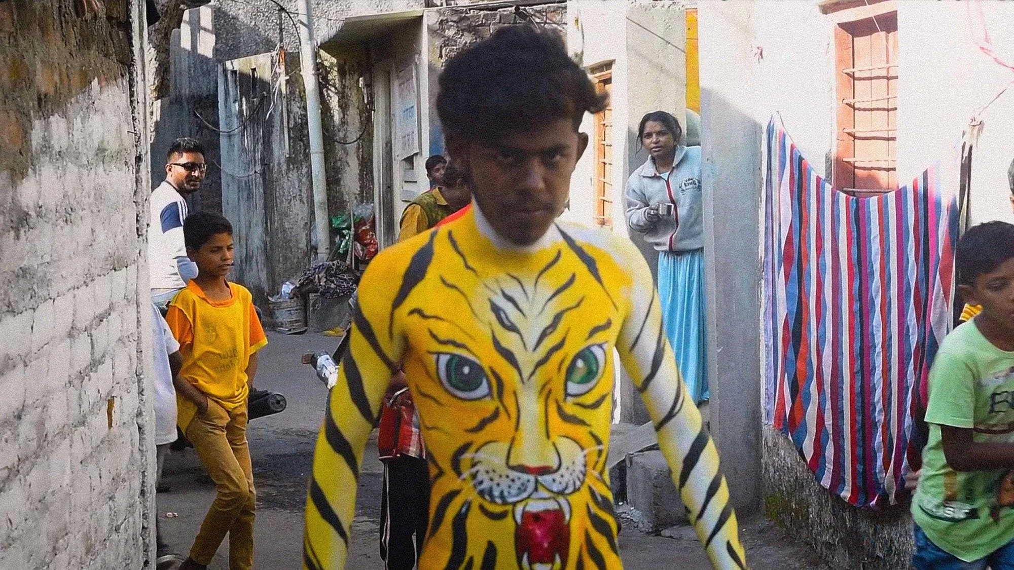 A young man walking down a street with a tiger face painted on his chest, surrounded by other people, some wearing colorful clothing, in a neighborhood with brick and concrete walls.