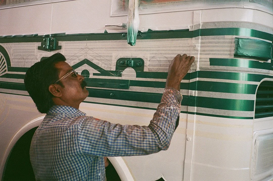 A man wearing glasses and a checkered shirt painting a green design on a white bus.