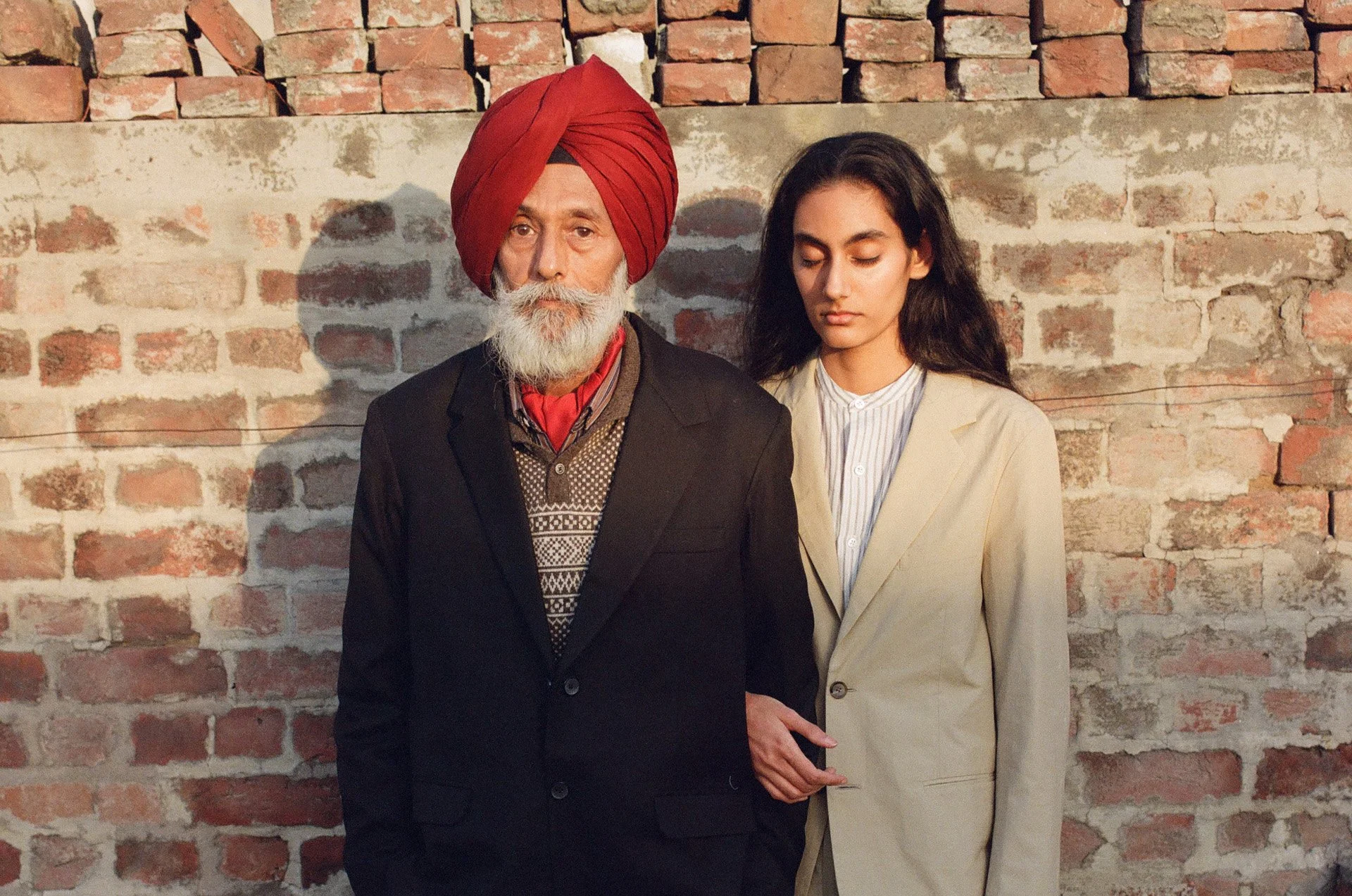 An older man with a white beard wearing a red turban and black suit, standing next to a young woman with long dark hair wearing a light-colored blazer and striped shirt, both standing against a brick wall with shadows cast behind them.