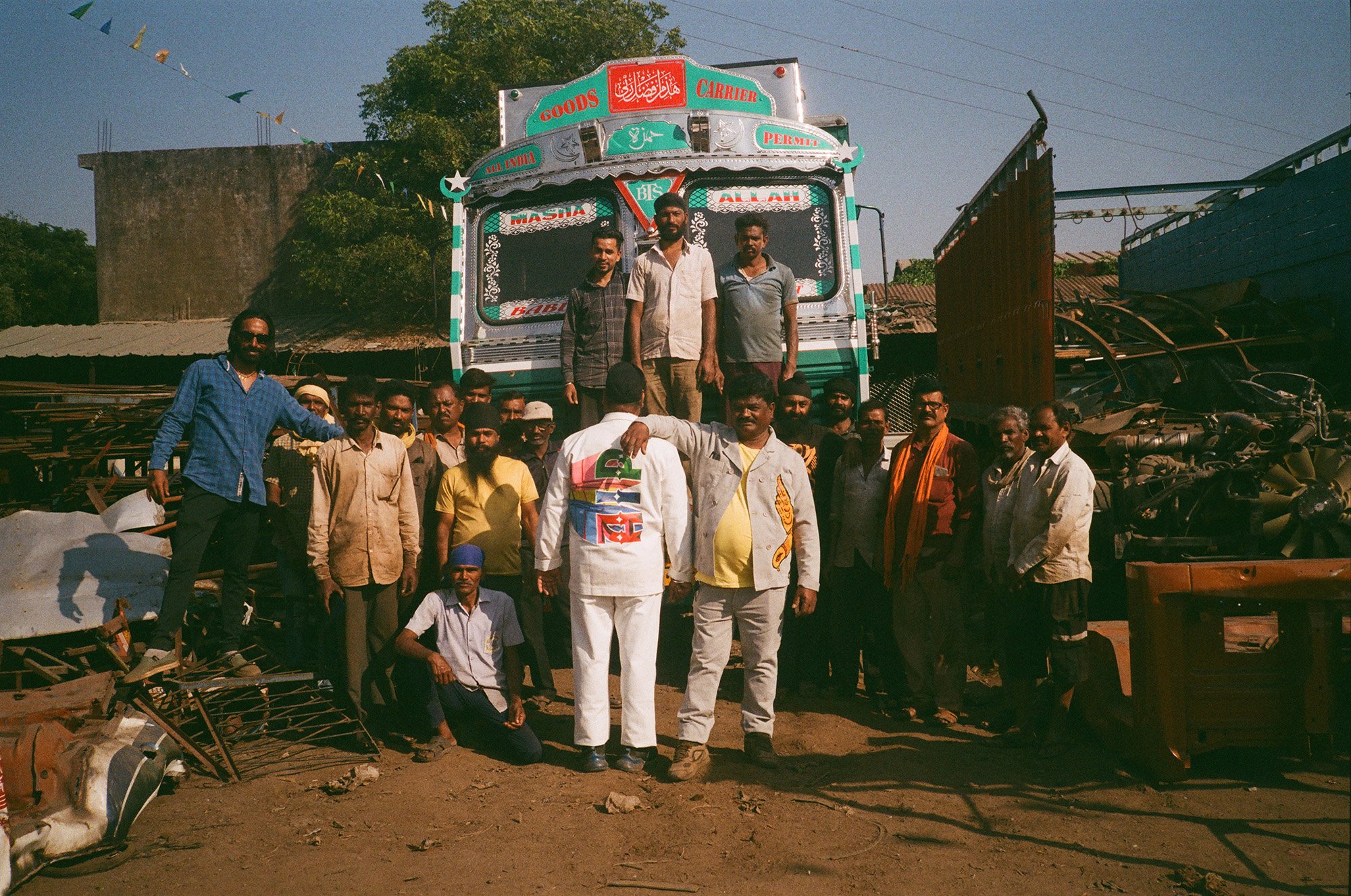 A group of men standing in front of a colorful truck, some standing on the truck and others on the ground. The background shows a rural setting with trees, a building, and miscellaneous machinery. The men are dressed in casual and traditional clothing, with some wearing sunglasses and head coverings.