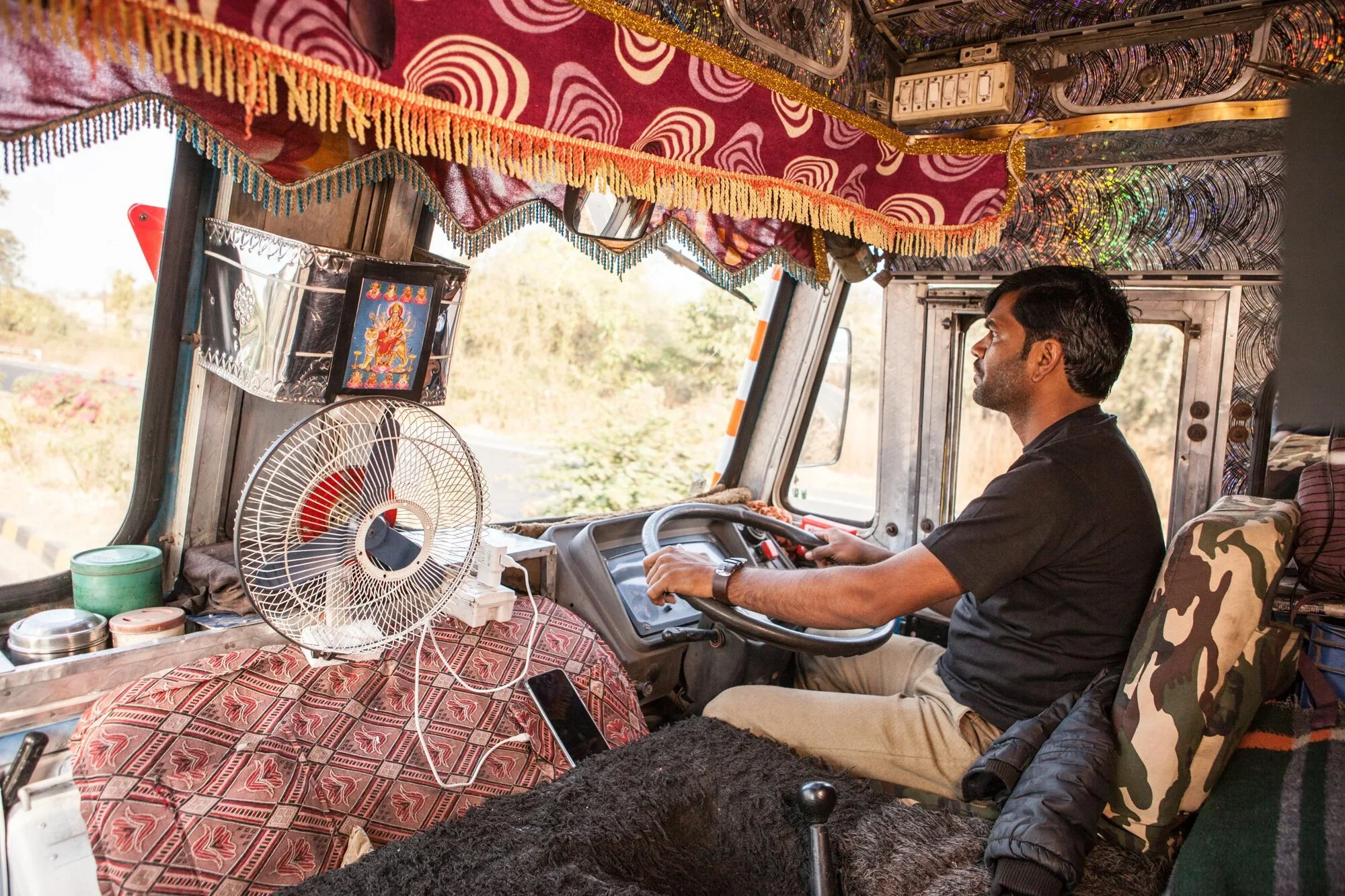 A man drives a truck decorated with colorful fabrics, fringe, and patterns, with an audience of prayer images and a fan on the dashboard.